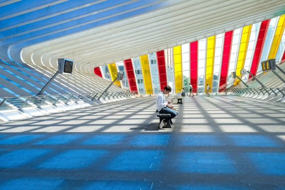 Person sitzt in einer modernen Halle mit bunten Streifen im Dach.