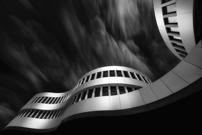 Moderne Architektur mit geschwungenen Linien und Fensterflächen vor dramatischem Himmel.