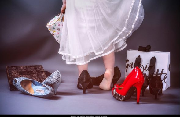Eine Person trägt schwarze Schuhe, umgeben von roten High Heels und verschiedenen Taschen.