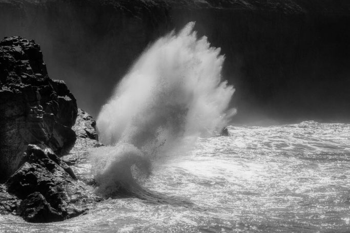 Spritzendes Wasser trifft auf Felsen am Ufer, monochrome Darstellung.