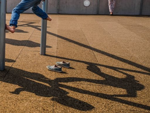 Ein Kind schwingt an einem Spielgerät, Schuhe liegen auf dem Boden, Schatten sind sichtbar.